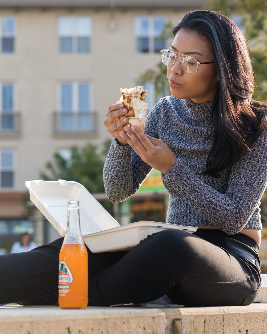 Customer eating a burrito outdoors with a Jarritos soda beside her.